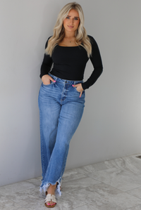 Woman wearing a black long sleeved fitted top tucked into high waisted distressed denim. The photo is taken indoors against a white wall with white marble flooring. 