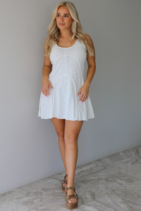 Woman wearing a white dress that has thick, tank-style straps, exposed seams details, has a lightweight t-shirt textured material, and has a mini dress length. The photo is taken indoors against a white wall with white marble flooring. 
