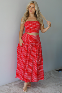 Woman wearing a red with white polka dots set featuring a cropped tank and high waisted maxi skirt indoors against a white wall with white marble flooring. 