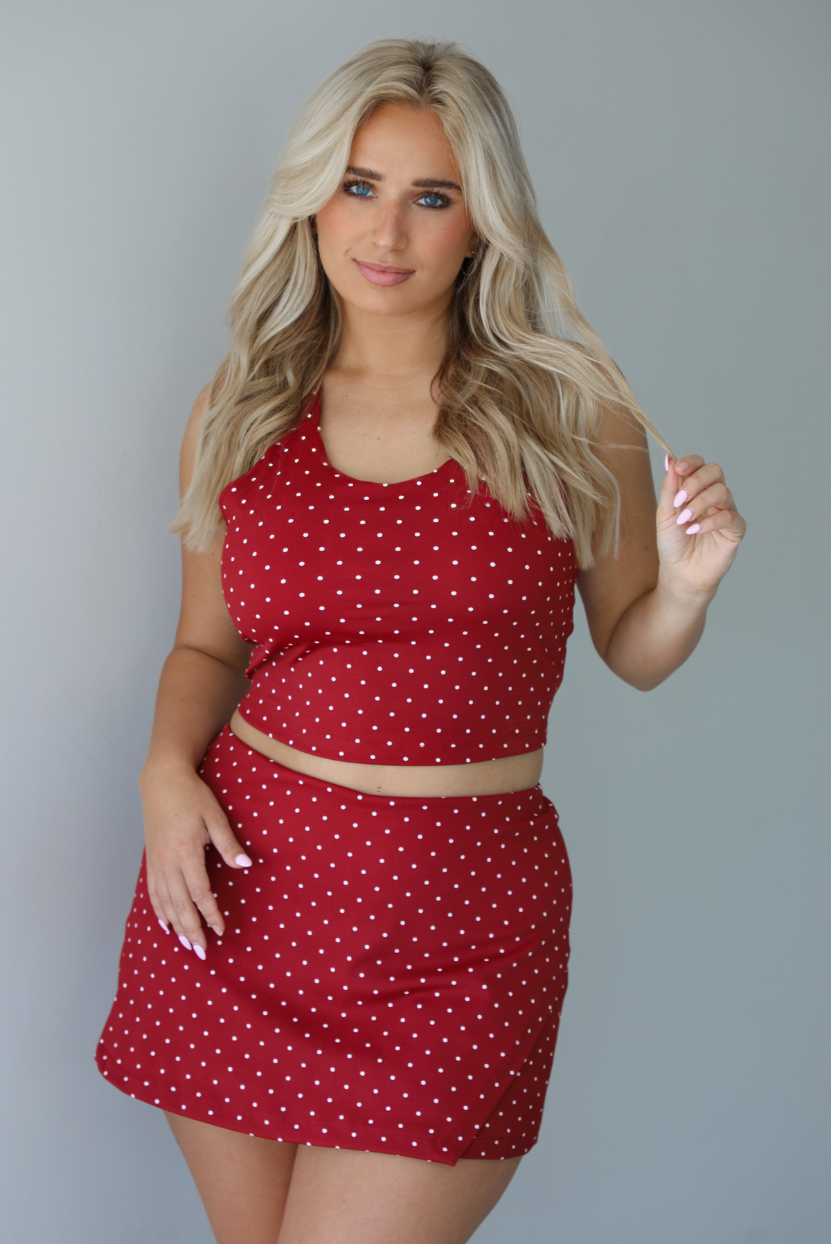 Woman wearing a red athleisure set with a white a white polka dot pattern throughout with a cropped tank and mini length skirt indoors against a white wall.