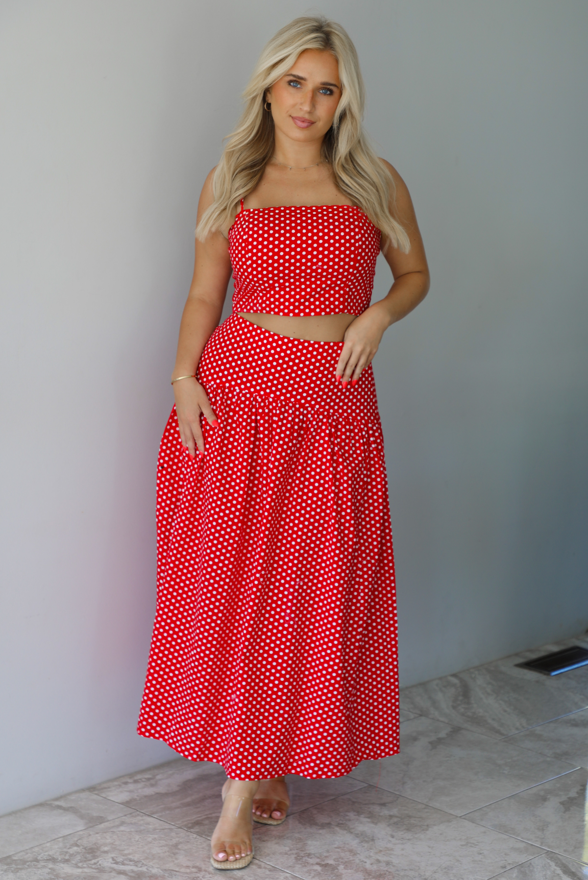 Woman wearing a red with white polka dots set featuring a cropped tank and high waisted maxi skirt indoors against a white wall with white marble flooring. 