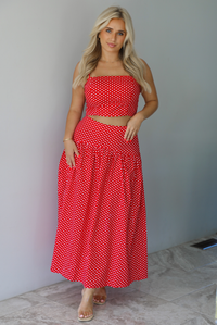 Woman wearing a red with white polka dots set featuring a cropped tank and high waisted maxi skirt indoors against a white wall with white marble flooring. 