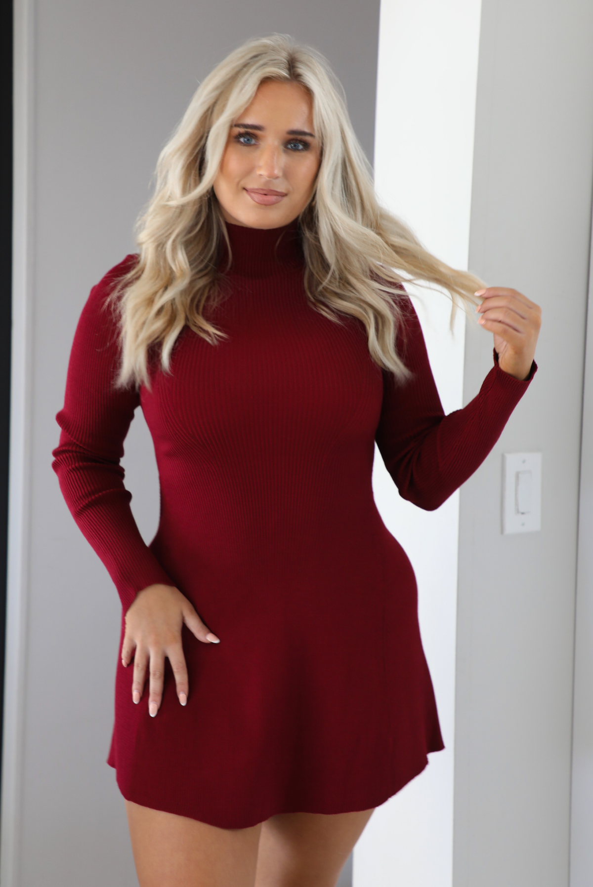 Woman model wearing a maroon colored sweater dress that has a high neckline, long sleeves, a ribbed texture, and stops on the upper thigh on the 5'7 model. The photo is taken indoors in an archway. The photo showcases a close up of the dress.