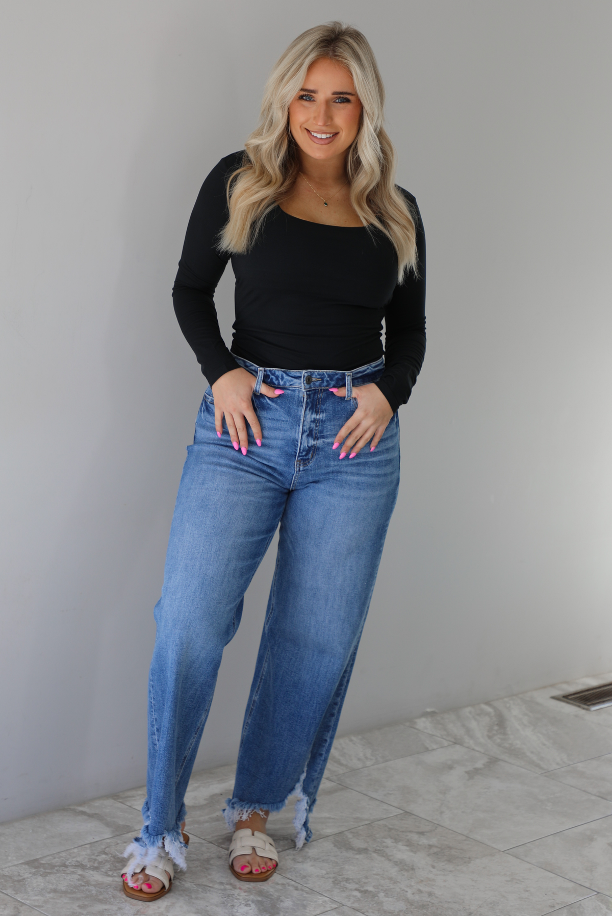 Woman wearing a black long sleeved fitted top tucked into high waisted distressed denim. The photo is taken indoors against a white wall with white marble flooring. 