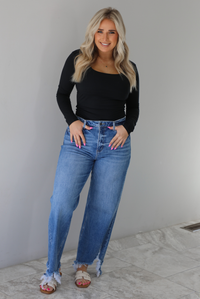 Woman wearing a black long sleeved fitted top tucked into high waisted distressed denim. The photo is taken indoors against a white wall with white marble flooring. 