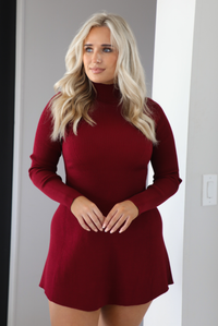 Woman model wearing a maroon colored sweater dress that has a high neckline, long sleeves, a ribbed texture, and stops on the upper thigh on the 5'7 model. The photo is taken indoors in an archway. The photo showcases a close up of the dress.