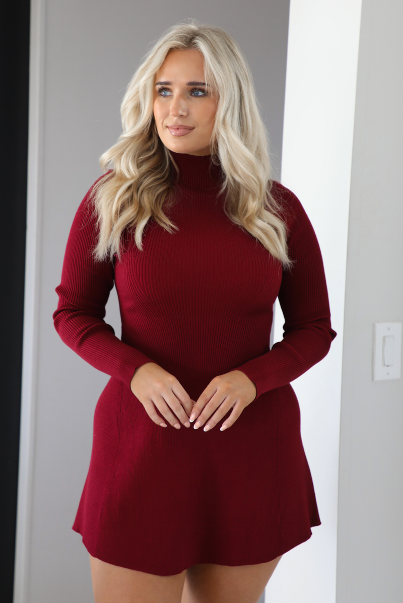 Woman model wearing a maroon colored sweater dress that has a high neckline, long sleeves, a ribbed texture, and stops on the upper thigh on the 5'7 model. The photo is taken indoors in an archway. The photo showcases a close up of the dress.