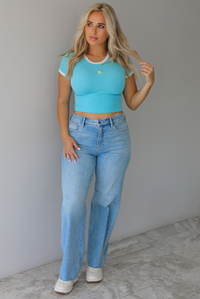 Woman wearing a blue crop top with an embroidered lemon on the front with mid-rise light-wash denim jeans against a white wall with white marble flooring. 