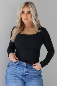 Woman wearing a black long sleeved fitted top untucked with high waisted distressed denim. The photo is taken indoors against a white wall.
