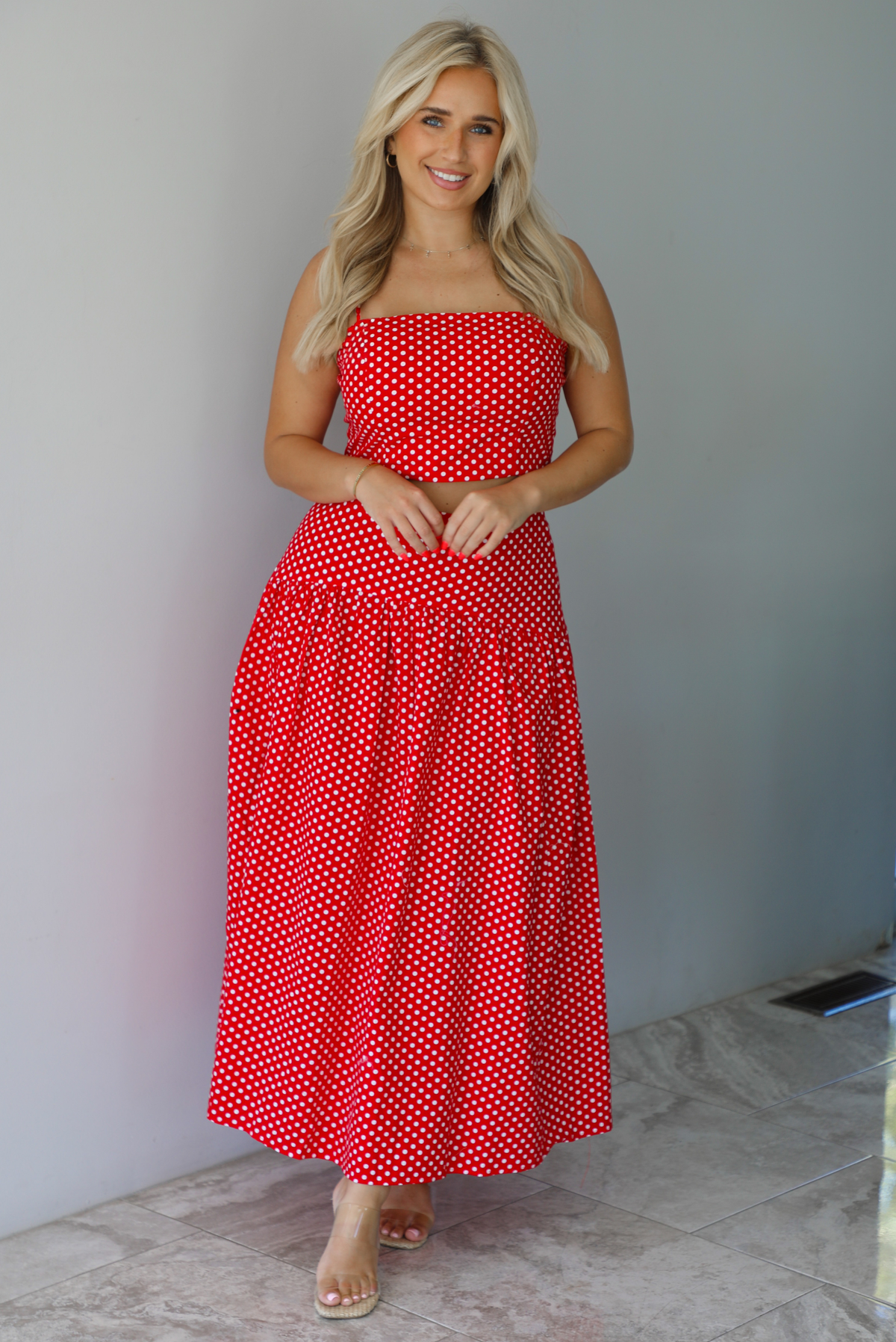 Woman wearing a red with white polka dots set featuring a cropped tank and high waisted maxi skirt indoors against a white wall with white marble flooring. 