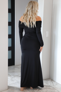 Woman wearing a fitted, black maxi dress that has a fold over detail at the shoulders, long sleeves, and a mermaid style skirt that's fitted at the hips and flows at the hem. The photo is worn indoors in a modern white home in an archway. The photo shows the back of the dress.