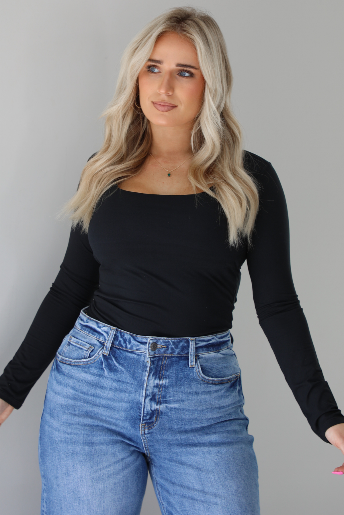 Woman wearing a black long sleeved fitted top tucked into high waisted distressed denim. The photo is taken indoors against a white wall.