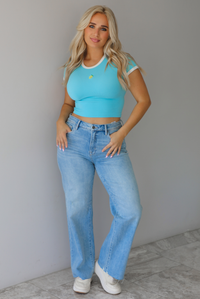 Woman wearing a blue crop top with an embroidered lemon on the front with mid-rise light-wash denim jeans against a white wall with white marble flooring. 