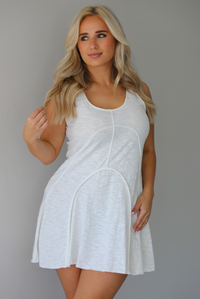 Woman wearing a white dress that has thick, tank-style straps, exposed seams details, has a lightweight t-shirt textured material, and has a mini dress length. The photo is taken indoors against a white wall.