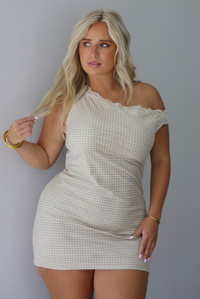 A woman wearing a cream and taupe plaid mini dress featuring one on the shoulder and one off the shoulder strap with lace detailing at the top hem of the dress, standing indoors against a white wall.