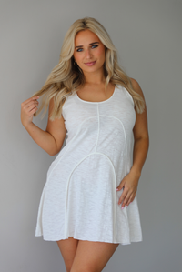 Woman wearing a white dress that has thick, tank-style straps, exposed seams details, has a lightweight t-shirt textured material, and has a mini dress length. The photo is taken indoors against a white wall.