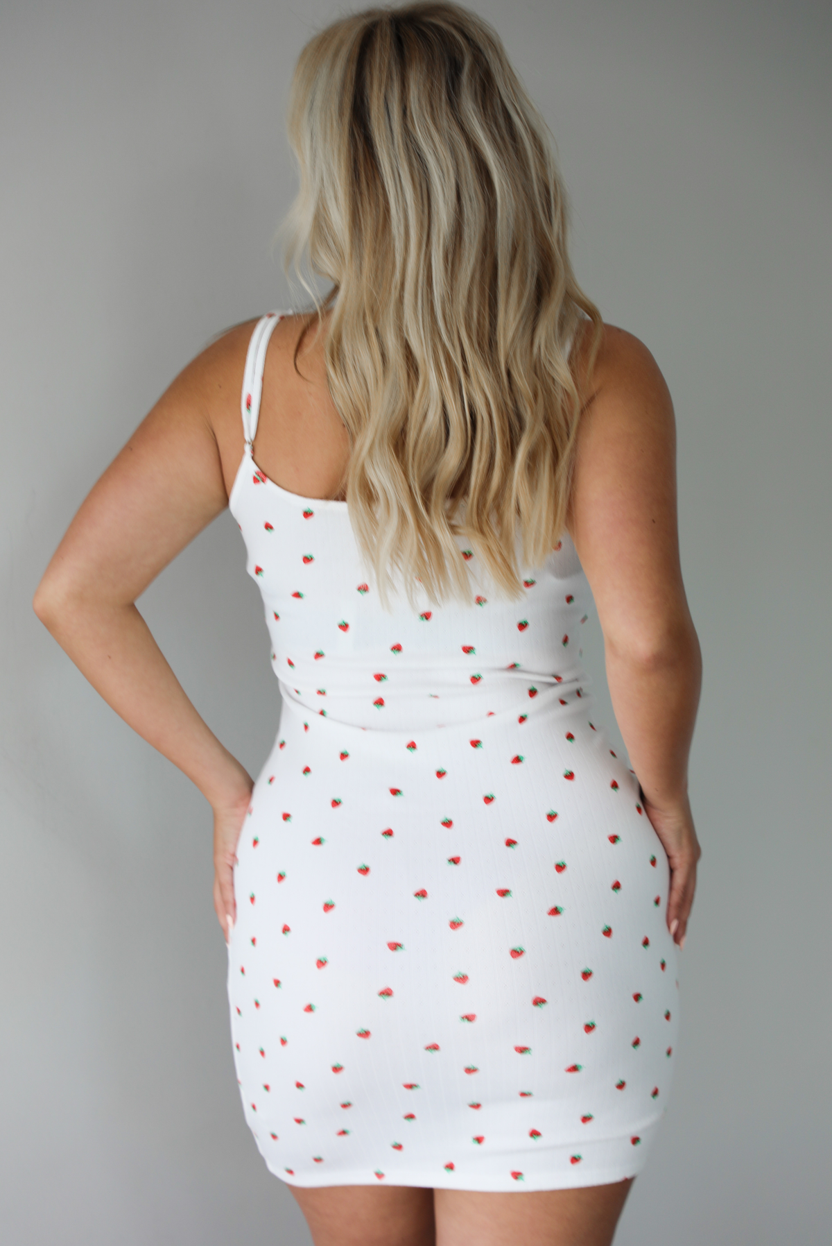 Woman wearing a white mini dress with a strawberry pattern indoors against a white wall. The photo shows the back of the dress.