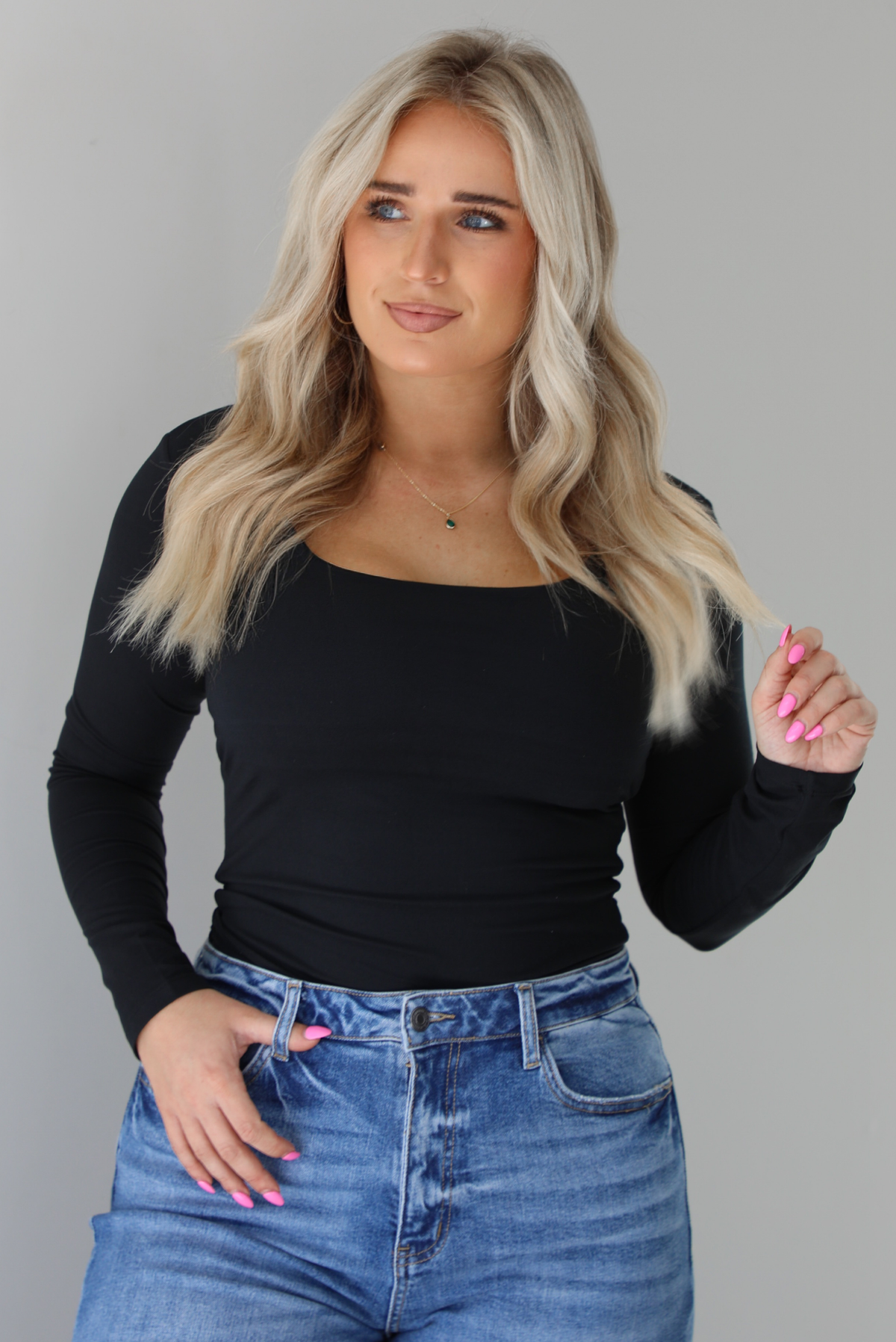 Woman wearing a black long sleeved fitted top tucked into high waisted distressed denim. The photo is taken indoors against a white wall.