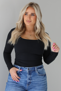 Woman wearing a black long sleeved fitted top tucked into high waisted distressed denim. The photo is taken indoors against a white wall.
