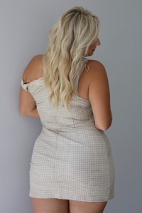 A woman shown from behind wearing a cream and taupe plaid mini dress featuring one on the shoulder and one off the shoulder strap with lace detailing at the top hem of the dress, standing indoors against a white wall.