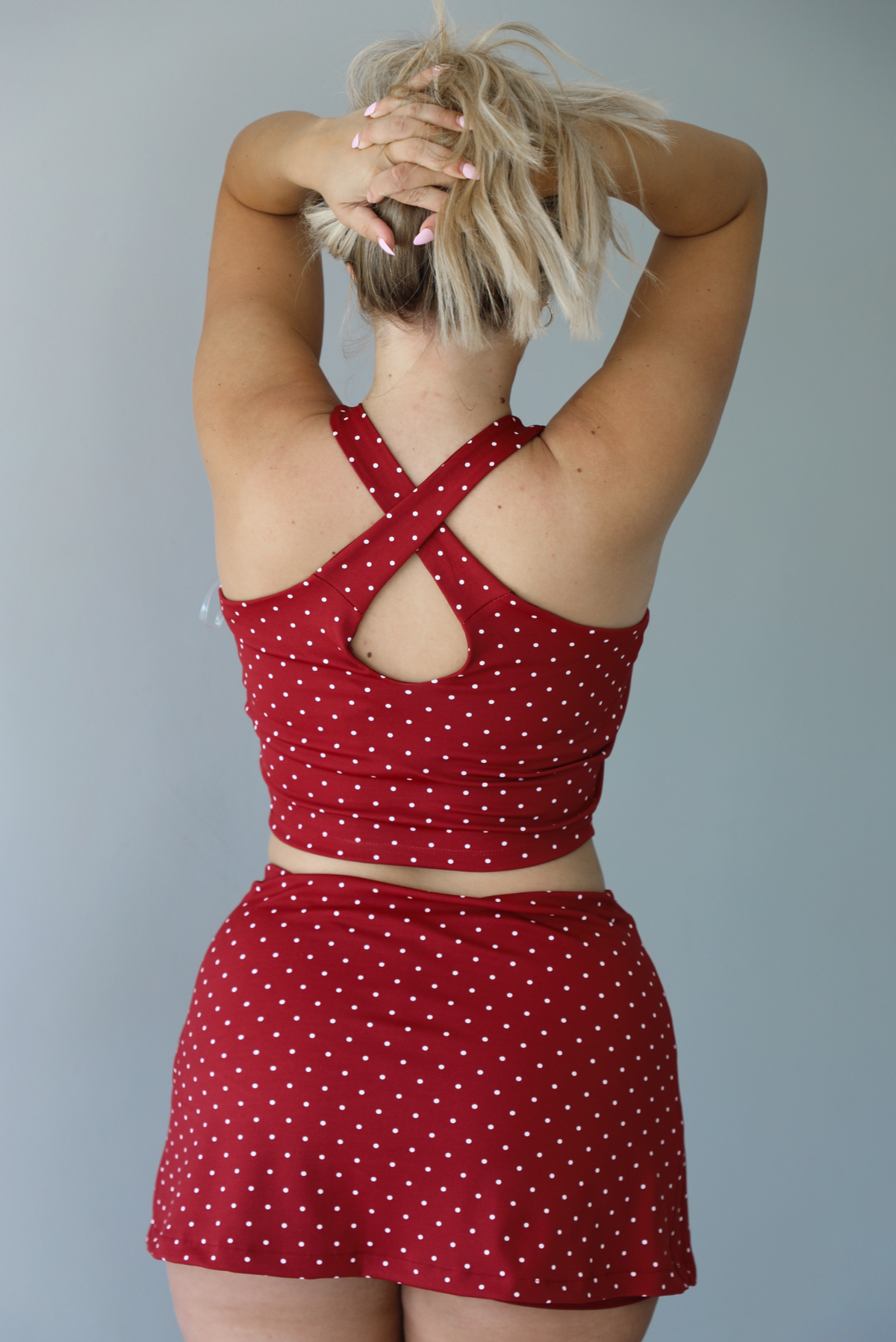 Woman wearing a red athleisure set with a white a white polka dot pattern throughout with a cropped tank and mini length skirt indoors against a white wall and shows the back of the set. 