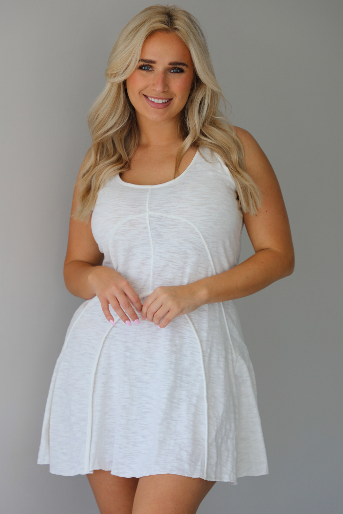 Woman wearing a white dress that has thick, tank-style straps, exposed seams details, has a lightweight t-shirt textured material, and has a mini dress length. The photo is taken indoors against a white wall.