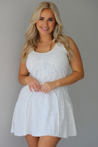 Woman wearing a white dress that has thick, tank-style straps, exposed seams details, has a lightweight t-shirt textured material, and has a mini dress length. The photo is taken indoors against a white wall.