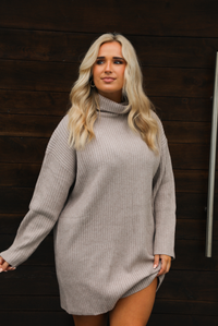 Woman wearing a taupe, ribbed sweater dress that has a slouched turtle neckline, long sleeves, and a relaxed fit. The photo is taken outdoors against a dark brown wooden door.
