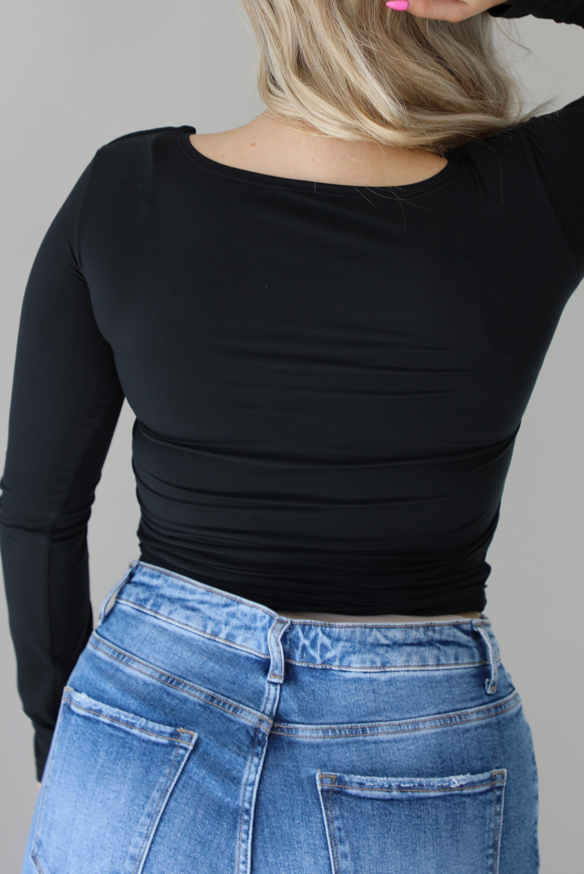 Woman wearing a black long sleeved fitted top untucked with high waisted distressed denim. The photo show the back of the top. The photo is taken indoors against a white wall.