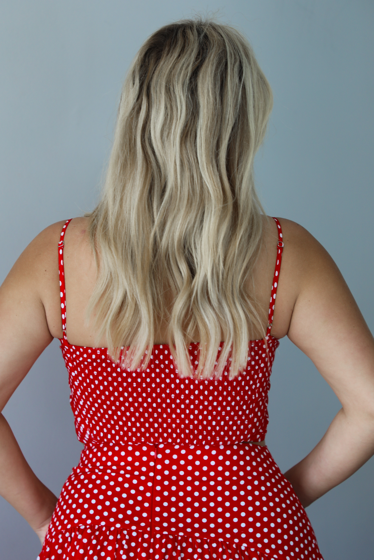 Woman wearing a red with white polka dots set featuring a cropped tank and high waisted maxi skirt indoors against a white wall. The photo shows the back of the tank, which has a smocked design.