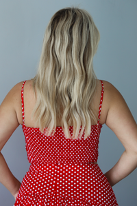 Woman wearing a red with white polka dots set featuring a cropped tank and high waisted maxi skirt indoors against a white wall. The photo shows the back of the tank, which has a smocked design.