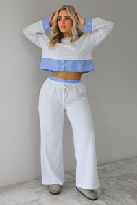 Woman wearing a heather grey two piece set with blue and white pin striped detailing at the hem of the top, at the wrists on the top, and at the waistline on the pants. The photo is taken indoors against a white wall with white marble flooring. 