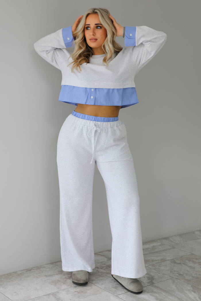 Woman wearing a heather grey two piece set with blue and white pin striped detailing at the hem of the top, at the wrists on the top, and at the waistline on the pants. The photo is taken indoors against a white wall with white marble flooring. 