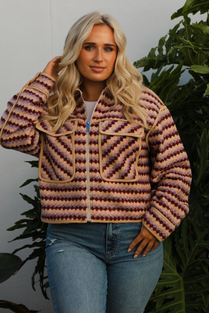 Jacket with a multi-colored sideways checkered pattern throughout, collared neckline, functional pockets in the front, and a functional zipper closure in the font. The jacket is paired with medium wash denim jeans and tan colored uggs. The photo is taken outdoors against a white wall with greenery in the back. The photo showcases the jacket close up.