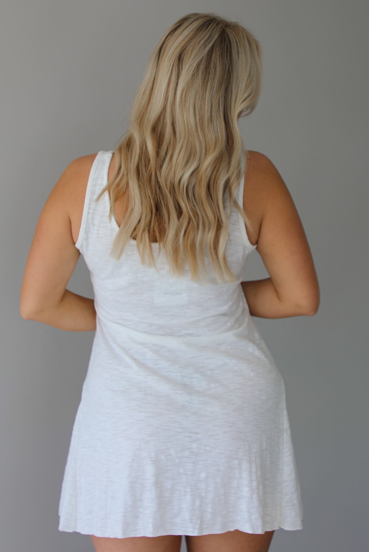 Woman wearing a white dress that has thick, tank-style straps, exposed seams details, has a lightweight t-shirt textured material, and has a mini dress length. The photo is taken indoors against a white wall and shows the back of the dress. 