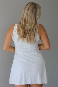 Woman wearing a white dress that has thick, tank-style straps, exposed seams details, has a lightweight t-shirt textured material, and has a mini dress length. The photo is taken indoors against a white wall and shows the back of the dress. 