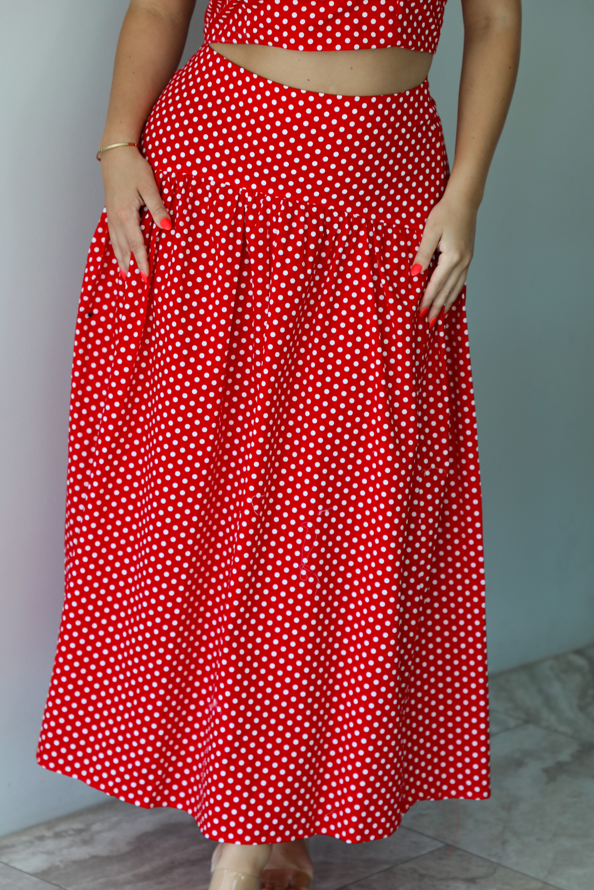 Woman wearing a red with white polka dots set featuring a cropped tank and high waisted maxi skirt indoors against a white wall with white marble flooring. The photo shows a closeup of the skirt. 
