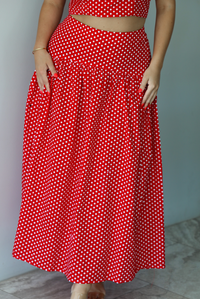 Woman wearing a red with white polka dots set featuring a cropped tank and high waisted maxi skirt indoors against a white wall with white marble flooring. The photo shows a closeup of the skirt. 