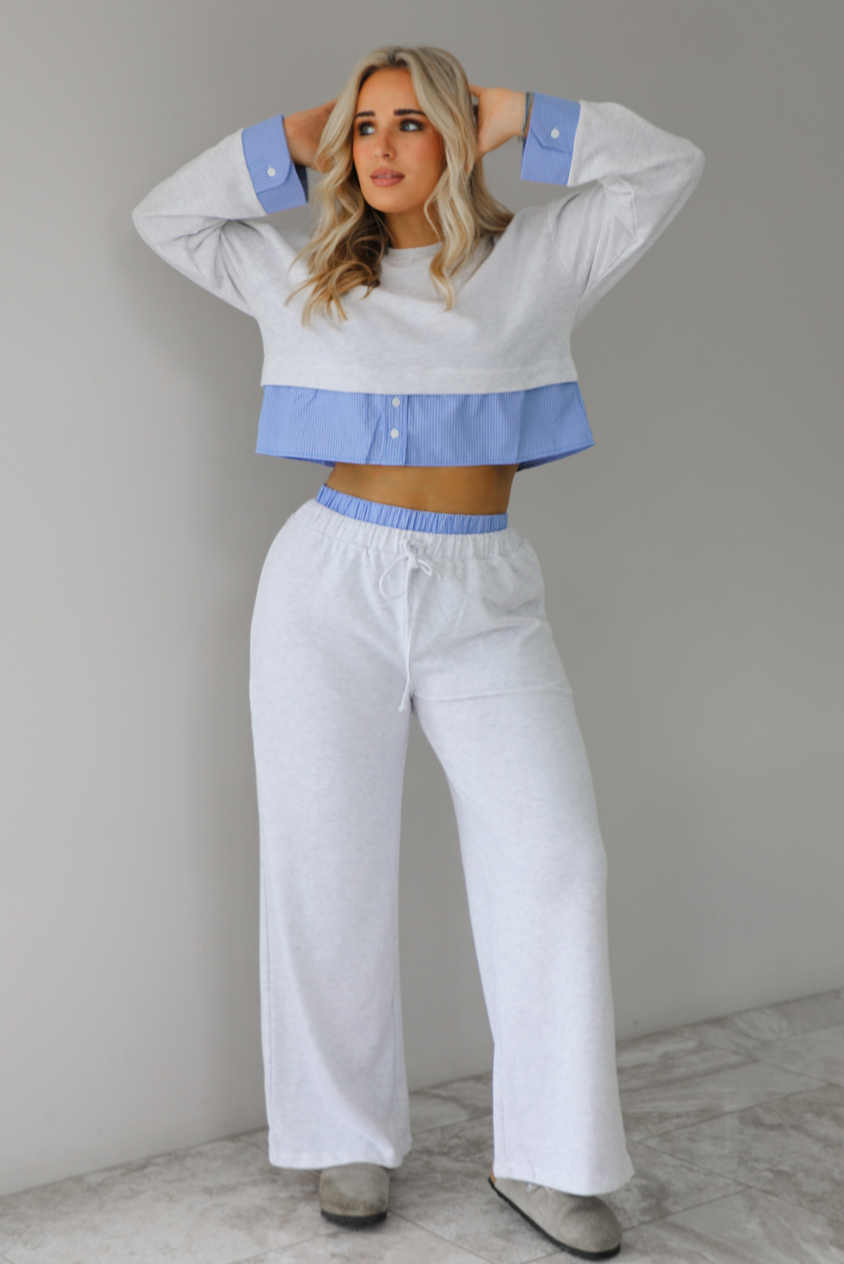 Woman wearing a heather grey two piece set with blue and white pin striped detailing at the hem of the top, at the wrists on the top, and at the waistline on the pants. The photo is taken indoors against a white wall with white marble flooring. 
