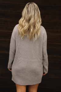 Woman wearing a taupe, ribbed sweater dress that has a slouched turtle neckline, long sleeves, and a relaxed fit. The photo is taken outdoors against a dark brown wooden door. The photo shows the back of the dress.