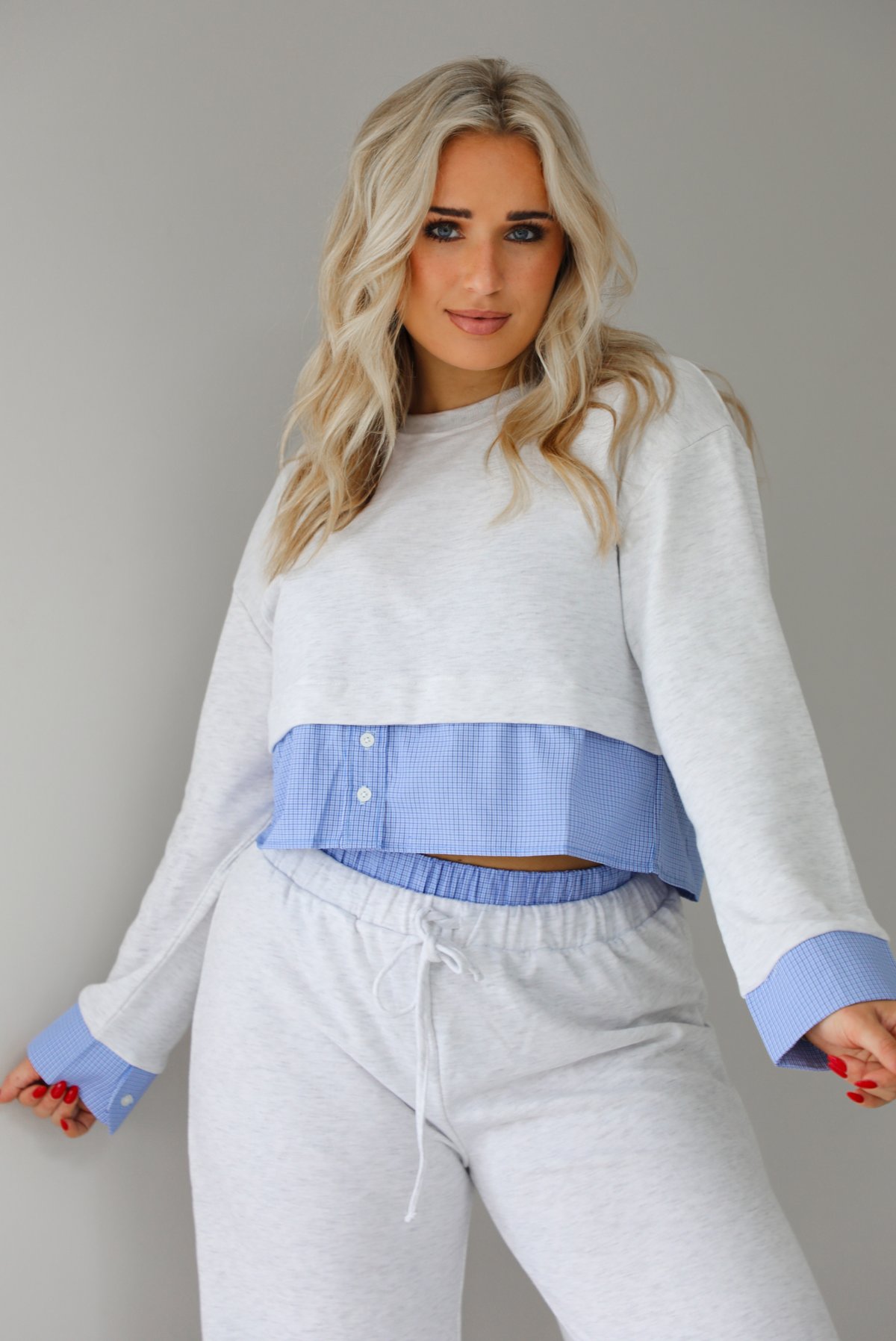 Woman wearing a heather grey two piece set with blue and white pin striped detailing at the hem of the top, at the wrists on the top, and at the waistline on the pants. The photo shows a closeup of the top and upper portion of the pants. The photo is taken indoors against a white wall with white marble flooring. 