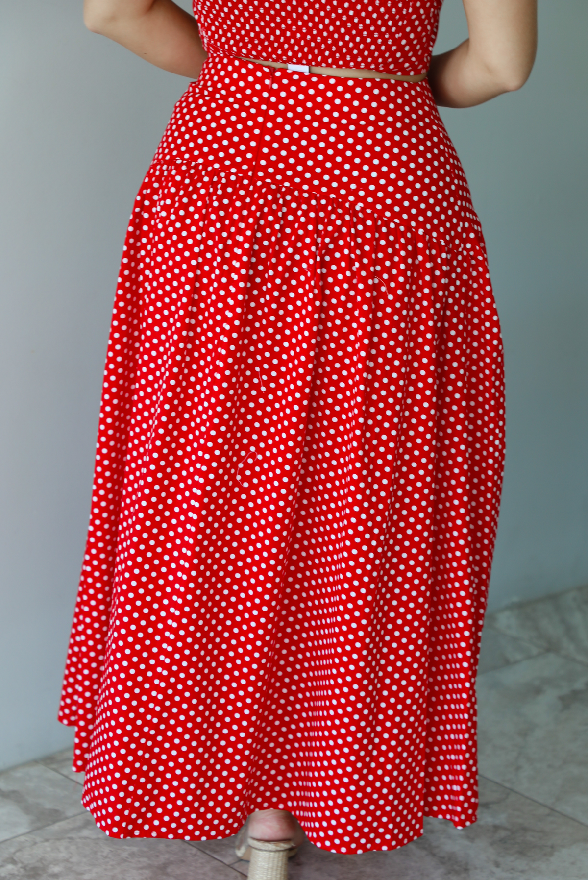 Woman wearing a red with white polka dots set featuring a cropped tank and high waisted maxi skirt indoors against a white wall with white marble flooring. The photo shows a closeup of the back of the skirt, which has a zipper closure. 