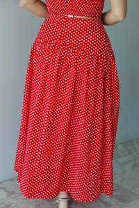 Woman wearing a red with white polka dots set featuring a cropped tank and high waisted maxi skirt indoors against a white wall with white marble flooring. The photo shows a closeup of the back of the skirt, which has a zipper closure. 