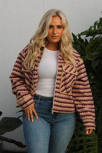 Jacket with a multi-colored sideways checkered pattern throughout, collared neckline, functional pockets in the front, and a functional zipper closure in the font. The jacket is paired with medium wash denim jeans and tan colored uggs. The photo is taken outdoors against a white wall with greenery in the back. The photo showcases the jacket close up unzipped.