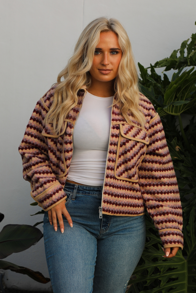 Jacket with a multi-colored sideways checkered pattern throughout, collared neckline, functional pockets in the front, and a functional zipper closure in the font. The jacket is paired with medium wash denim jeans and tan colored uggs. The photo is taken outdoors against a white wall with greenery in the back. The photo showcases the jacket close up unzipped.