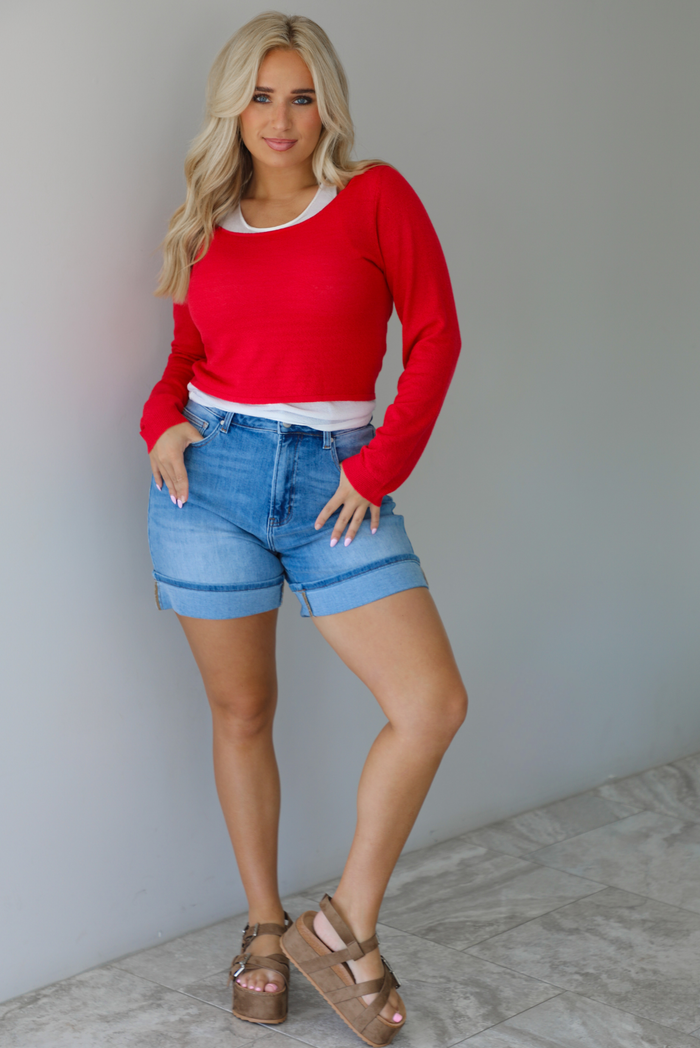 Woman wearing a red, long sleeved knit top with a white tank underneath with medium wash denim shorts indoors against a white wall with white marble flooring. 
