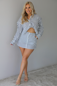 Woman wearing a white and navy striped set with a collared button-down top and mini skirt indoors against a white wall with white marble flooring. 