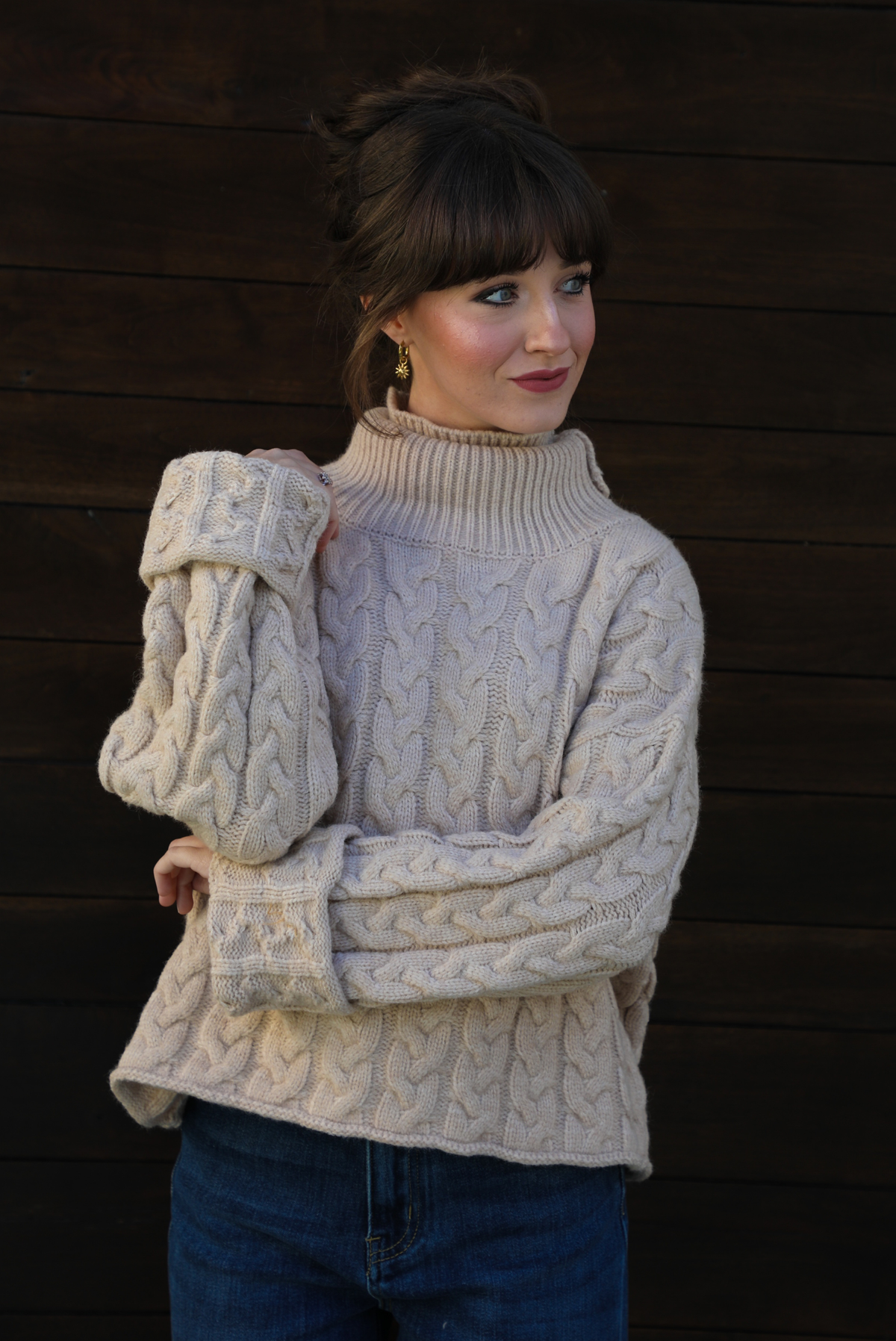 Woman model wearing a tan, thick, cable knit turtle neck sweater with dark denim jeans outdoors in front of a dark stained wooden door. 