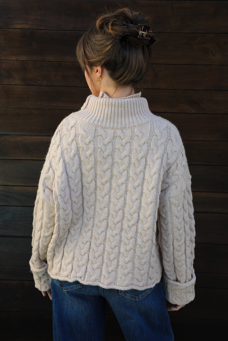 Woman model wearing a tan, thick, cable knit turtle neck sweater with dark denim jeans outdoors in front of a dark stained wooden door. 