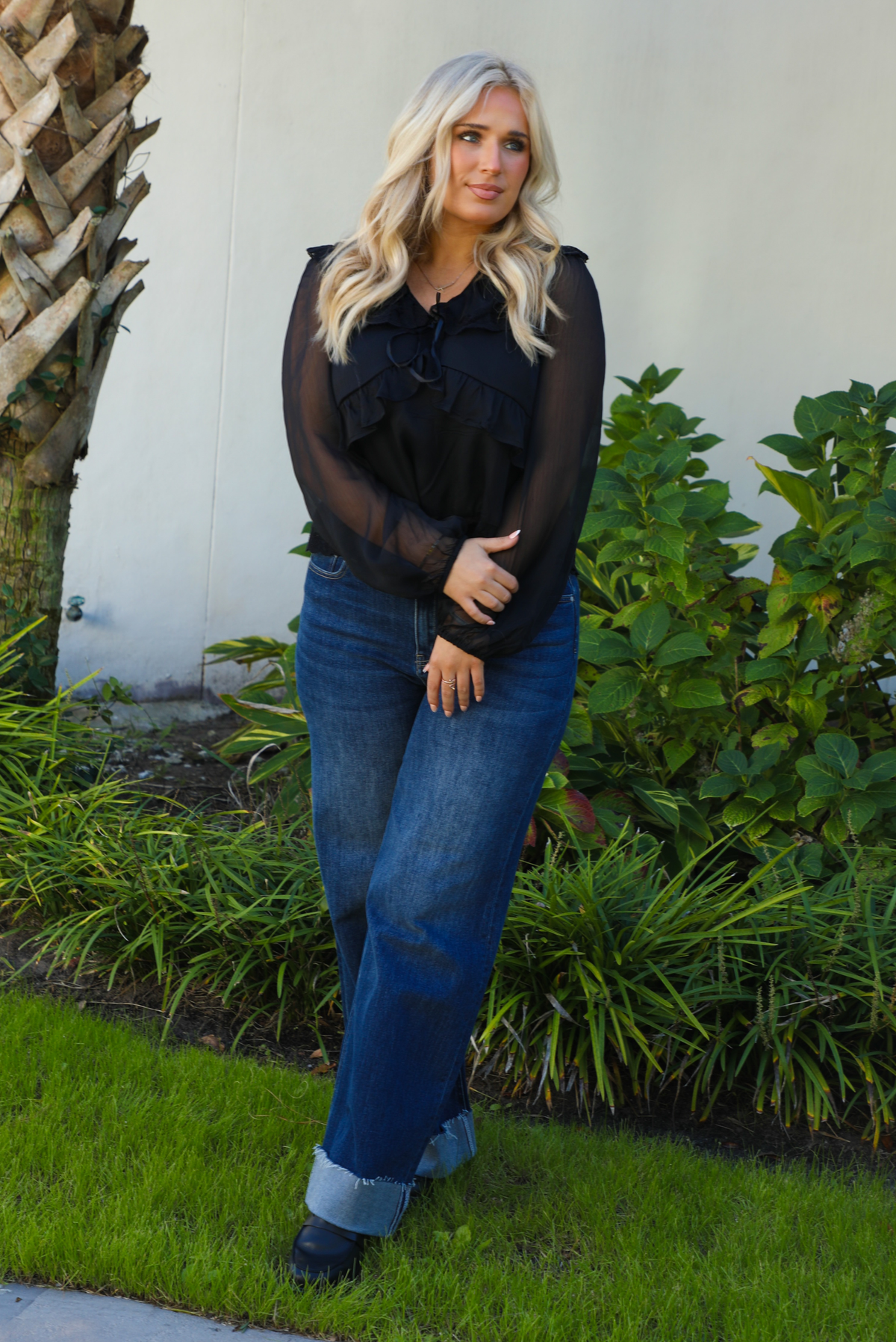 Woman model wearing a black top and blue jeans standing outdoors with greenery in the background.